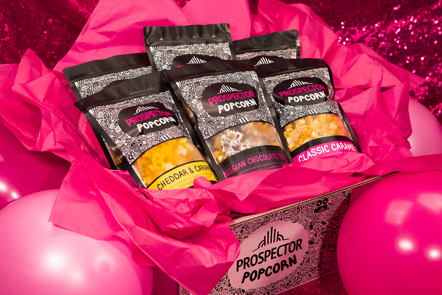 A gourmet popcorn gift box sits on a table surrounded by pink balloons and pink sparkle.