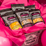 A gourmet popcorn gift box sits on a table surrounded by pink balloons and pink sparkle.