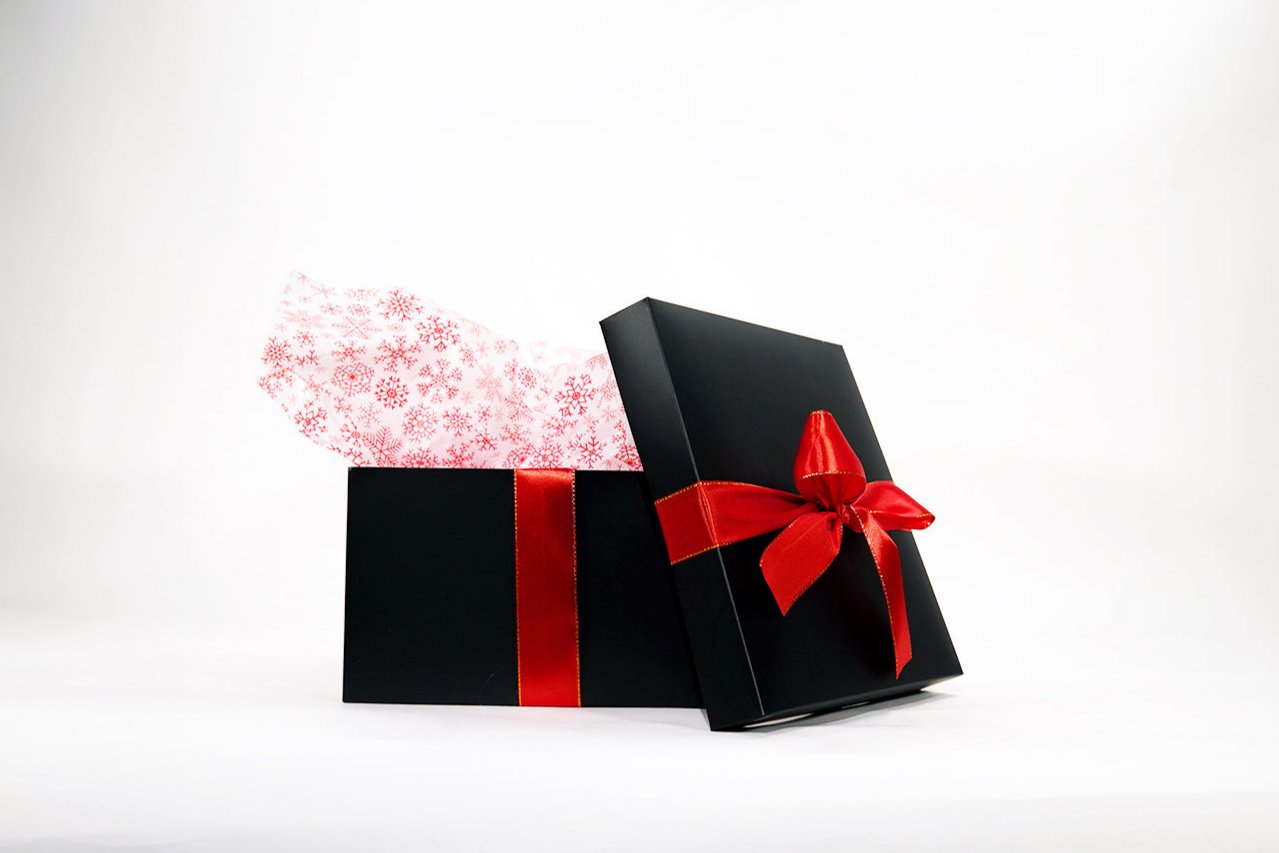 A black gift box with a red ribbon is partially open, revealing white tissue paper with red snowflake patterns against a white background.