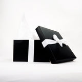 A black gift box with a white ribbon is partially open, showing white tissue paper. The box is set against a plain white background.