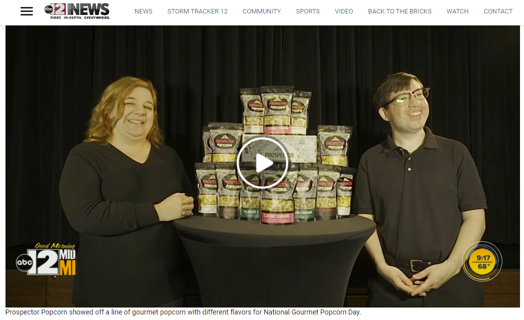 Two employees of prospector popcorn dressed in black show prospector popcorn bags to camera for live tv appearance