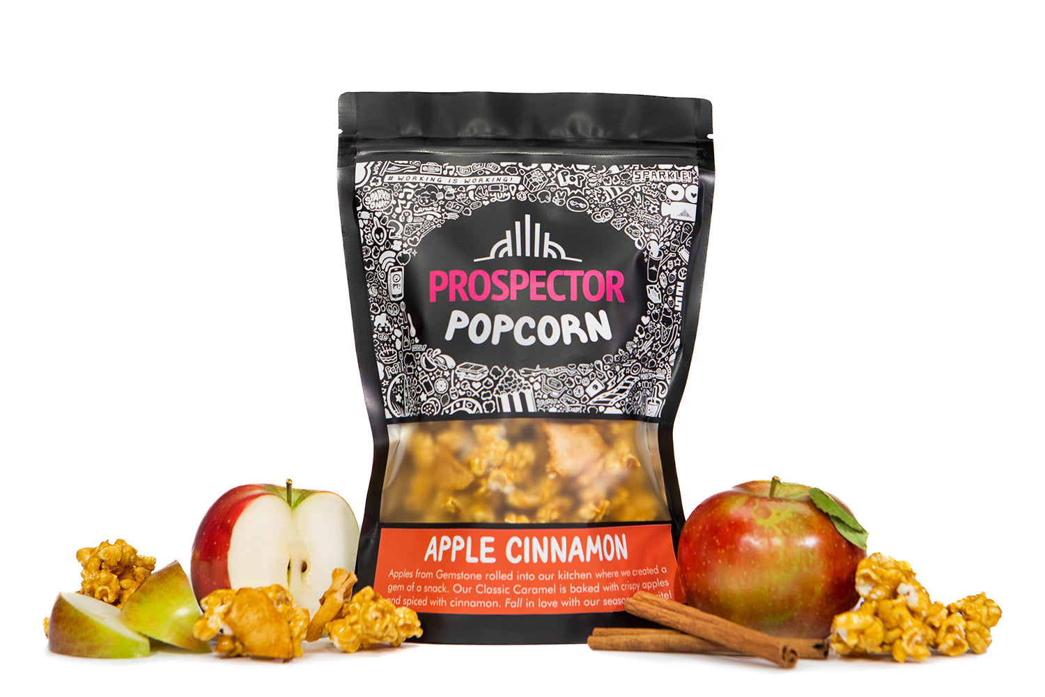 A bag of Apple Cinnamon Prospector gourmet popcorn sits next to apples, sliced and whole, cinnamon sticks, and popcorn.