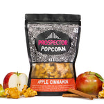 A bag of Apple Cinnamon Prospector gourmet popcorn sits next to apples, sliced and whole, cinnamon sticks, and popcorn.