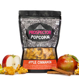 A bag of Apple Cinnamon Prospector gourmet popcorn sits next to apples, sliced and whole, cinnamon sticks, and popcorn.