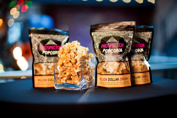 Three bags of Million Dollar Baby gourmet popcorn on a black table cloth. A small square glass jar vase with gourmet popcorn is filled and is placed with the three bags