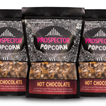 Three bags of Prospector Popcorn, labeled "Hot Chocolate" flavor, are lined up. The packaging features intricate black and white patterns with a clear window showing the popcorn inside. Each bag includes text about the hot chocolate flavor.