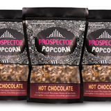 Three bags of Prospector Popcorn, labeled "Hot Chocolate" flavor, are lined up. The packaging features intricate black and white patterns with a clear window showing the popcorn inside. Each bag includes text about the hot chocolate flavor.