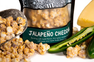 A bag of Jalapeno Cheddar gourmet popcorn sits behind its popcorn, a scooper filled with popcorn, jalapeños, and cheddar cheese wedges.