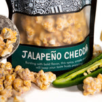 A bag of Jalapeno Cheddar gourmet popcorn sits behind its popcorn, a scooper filled with popcorn, jalapeños, and cheddar cheese wedges.