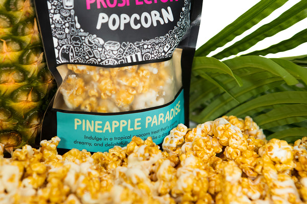 A bag of Pineapple Paradise popcorn sits in a pile of its popcorn. A pineapple and palm leaves sit behind it