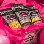 A gourmet popcorn gift box sits on a table surrounded by pink balloons and pink sparkle.