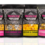 Eight bags of Prospector Popcorn is a row with eight different flavors on a white background
