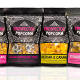 Eight bags of Prospector Popcorn is a row with eight different flavors on a white background