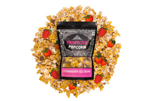 A bag of Strawberry Ice Cream Gourmet Popcorn lays on a pile of its popcorn. 4 strawberries lay on the pile around the bag.