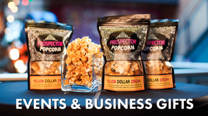 Three bags of Million Dollar Baby gourmet popcorn on a black table cloth. A small square glass jar vase with gourmet popcorn is filled and is placed with the three bags