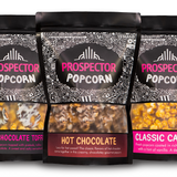 Three bags of Prospector Popcorn are displayed. Flavors include Belgian Chocolate Toffee, Hot Chocolate, and Classic Caramel. Each bag features detailed decorative borders and clear panels showing the popcorn inside.