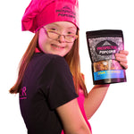 Popcorn Chef, Brooklyn, holds a bag of Down to Sparkle gourmet popcorn as she wears a pink apron and pink chefs hat.