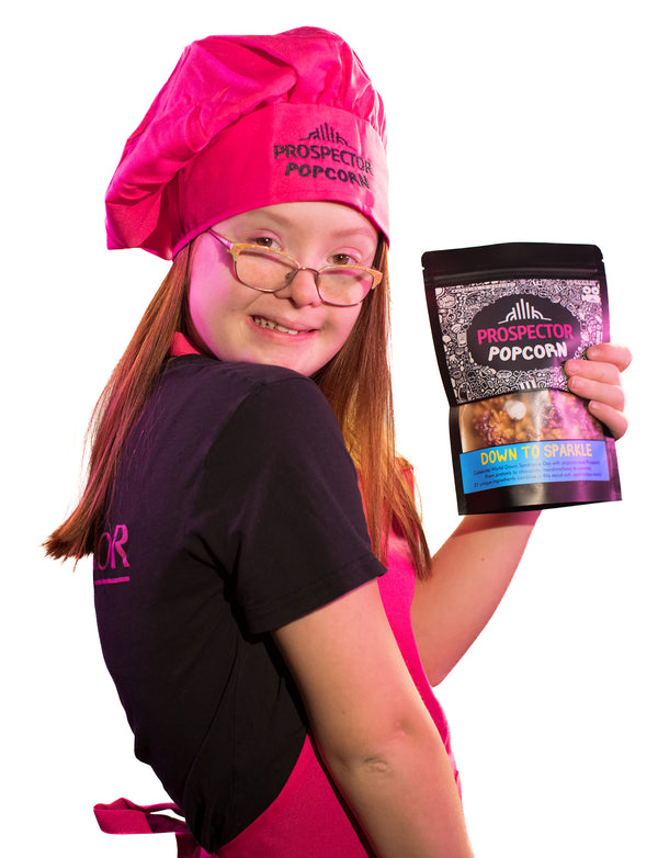 Popcorn Chef, Brooklyn, holds a bag of Down to Sparkle gourmet popcorn as she wears a pink apron and pink chefs hat.