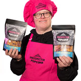 Popcorn Chef David holds two bags of Down to Sparkle gourmet popcorn as he wears a pink apron and pink chefs hat.