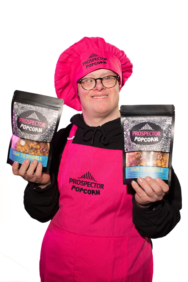Popcorn Chef David holds two bags of Down to Sparkle gourmet popcorn as he wears a pink apron and pink chefs hat.