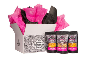 3 bags of 2oz gourmet popcorn bags sit in front of a white box printed with the Prospector Popcorn label on it. Pink and black tissue paper pops out of the top of the box. Buffalo Cheddar, Belgian Chocolate Toffee, Cheddar and Caramel are shown,