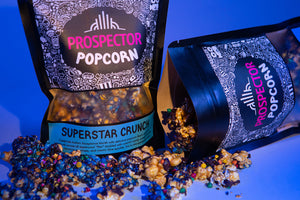 A bag of Superstar Crunch popcorn sits on a blue background as another bag has been spilled in front of it.